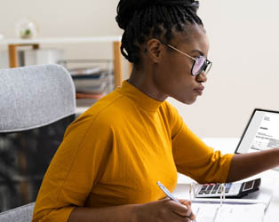 Imagem de uma jovem negra de óculos, sentada em uma mesa de perfil, em um fundo claro, usando uma blusa amarela, trabalhando em dois computadores em planilhas administrativas