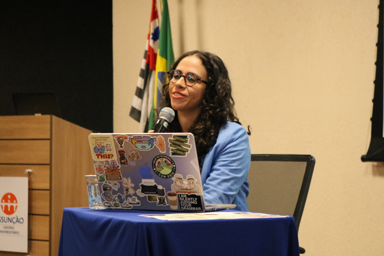 Professora Natália Mendes Teixeira sorri enquanto segura o microfone, sentada à mesa com notebook adesivado, com a bandeira do Brasil e de São Paulo ao fundo.