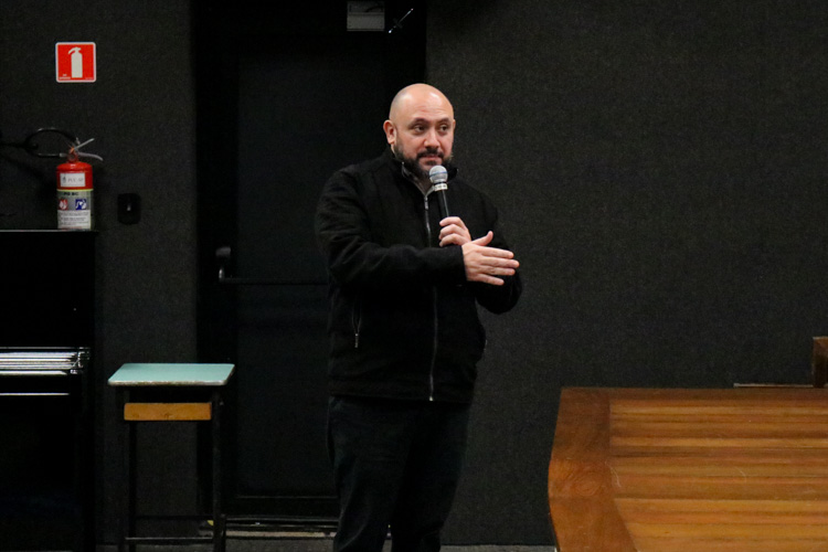 Professor faz uma intervenção de fala durante o evento, segurando um microfone em uma das mãos, posicionado à frente do palco no auditório.