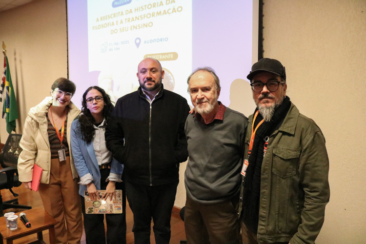 Registro em grupo com organizadores e palestrante: cinco pessoas posam sorrindo para a foto, em frente à tela de projeção da palestra.