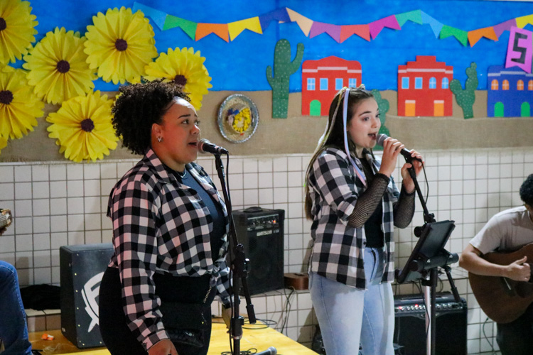 Duas cantoras, vestidas com camisas xadrez, garantindo música ao-vivo no arraiá do assunção