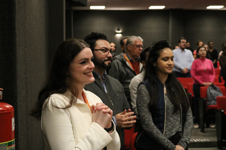 Mulher de branco, sorridente, com as mãos em oração, ao lado de outros participantes atentos na plateia da Vigília Universitária.