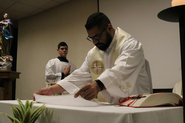 Padre de óculos e estola com símbolo do Espírito Santo organiza o altar, com acólito ao fundo e imagem de santo ao lado.