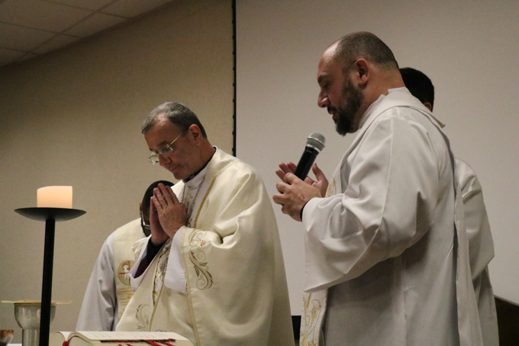 Dom Carlos, de cabeça baixa e mãos postas em oração, ao lado do Padre Rodrigo Pires que segura um microfone e também ora diante do altar iluminado por velas.