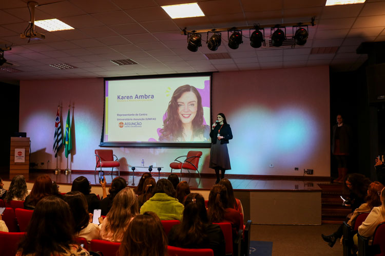Professora Karen Ambra fala no palco do auditório do Centro Universitário Assunção, com público assistindo e apresentação projetada ao fundo com seu nome e foto.