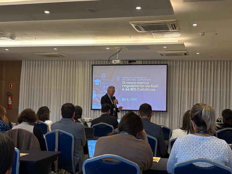 Palestrante em pé, com microfone na mão, fala a um auditório durante o Seminário Presencial da ANEC sobre o novo marco regulatório da EAD e as IES Católicas. Ao fundo, uma tela de projeção exibe o título do evento. Participantes estão sentados, de costas, atentos à apresentação.
