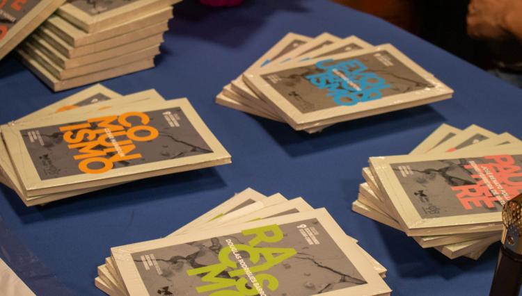 Livros da coleção 'Desconstruindo o Lugar-Comum' organizados sobre uma mesa com toalha azul. Entre os títulos visíveis estão 'Comunismo', 'Racismo', 'Paulo Freire' e 'Imperialismo', cada um com design gráfico destacado em cores vibrantes, como laranja, verde e azul