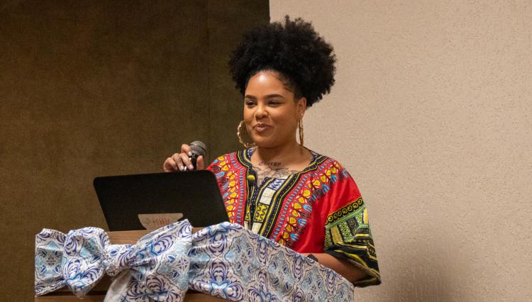 Mulher negra apresentando uma palestra, usando microfone e laptop. Ela veste uma blusa com estampa colorida de inspiração africana, com um sorriso confiante. Ao fundo, parede neutra e à frente, um púlpito decorado com tecido azul estampado e um laço grande