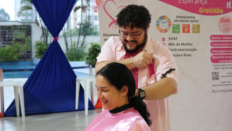 Um cabeleireiro sorridente, vestindo uma camisa rosa, corta uma mecha de cabelo de uma mulher que também veste um avental rosa e sorri. Ao fundo, há um banner do Instituto Amor em Mechas com palavras como 'amor', 'autoestima' e 'esperança', além de informações sobre a campanha de doação de cabelos