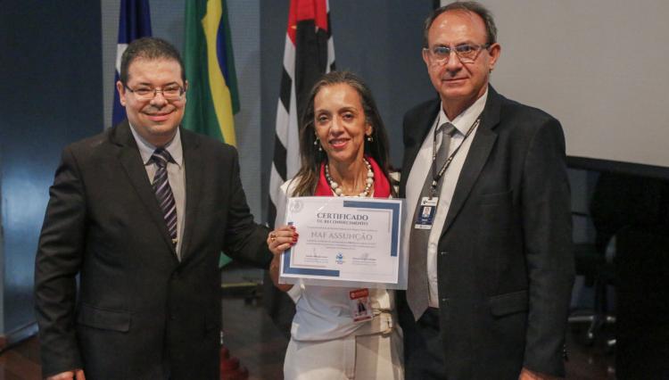 Três pessoas estão posando para uma foto em um evento formal. A mulher ao centro, vestindo roupa branca com detalhes vermelhos e um colar de pérolas, segura um certificado de reconhecimento intitulado "NAF ASSUNÇÃO". Ela sorri enquanto é acompanhada por dois homens, ambos usando ternos escuros e crachás. O homem à esquerda usa óculos e uma gravata listrada, enquanto o homem à direita também usa óculos e um crachá pendurado em um cordão.