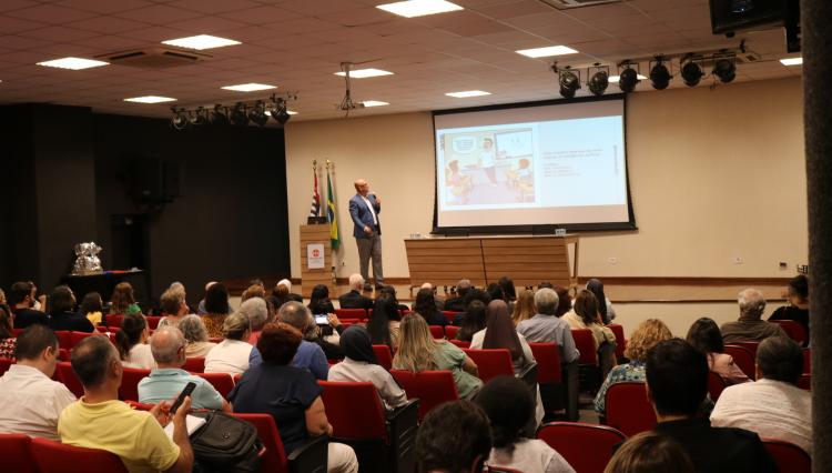 Auditório com público assistindo a uma palestra. No palco, um homem apresenta um slide sobre inteligência artificial na educação.