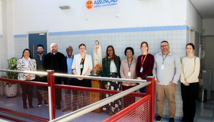 Membros da Secretaria Municipal de Direitos Humanos e Cidadania de São Paulo, ao lado da Reitora e representantes do Centro Universitário Assunção, posam para foto durante a visita institucional. O grupo está em um ambiente interno, com paredes de azulejos claros e o logotipo da instituição ao fundo.